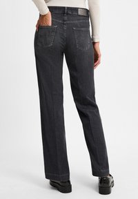 Dunkelgraue weite Jeans mit glatter Textur, ausgestattet mit zwei Gesäßtaschen und dezenten Stickdetails, getragen mit schwarzen Plateauschuhen.