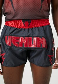 Schwarze Sportshorts mit einem roten elastischen Bund; verfügt über ein glänzendes rotes "VENUM"-Logo auf der Rückseite und Blitzakzente an den Seiten.