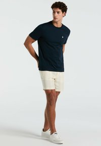 Marineblaues T-Shirt mit kurzen Ärmeln, einer Tasche und Logo, kombiniert mit hellbeigen gemusterten Shorts und weißen Sneakers. Glatte Stoffoberfläche.