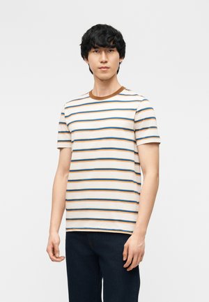 Homme portant un T-shirt crème à manches courtes avec des rayures horizontales bleues et marron, associé à un pantalon foncé, debout sur fond blanc.