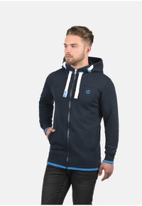 Marinefarbener Zip-Hoodie mit weißen Kordeln, blauen Akzenten und vorderen Taschen. Merkmale einen glatten Stofftextur und ein tailliertes Design.