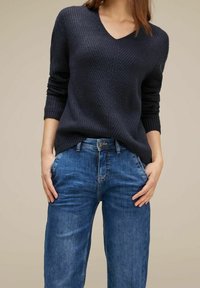 Pull en maille bleu marine avec col en V, manches longues et design texturé, associé à un jean denim bleu clair avec une coupe classique et des poches.
