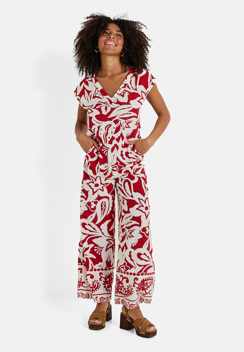 Mono floral rojo y blanco con mangas cortas, escote en V, dos bolsillos laterales y piernas de pantalón acampanadas. Llevado con zapatos de plataforma marrones.