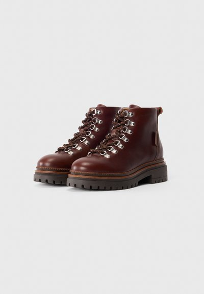 Brūnu ādu lace-up zābaku pāris ar biezām melnām zālēm un sudraba acs caurumiem uz balta fona.