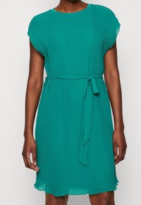 Tealfärgad plisserad klänning med korta ärmar, rund halsringning och ett tygbälte. Har en utsvängd fåll och subtil struktur.
