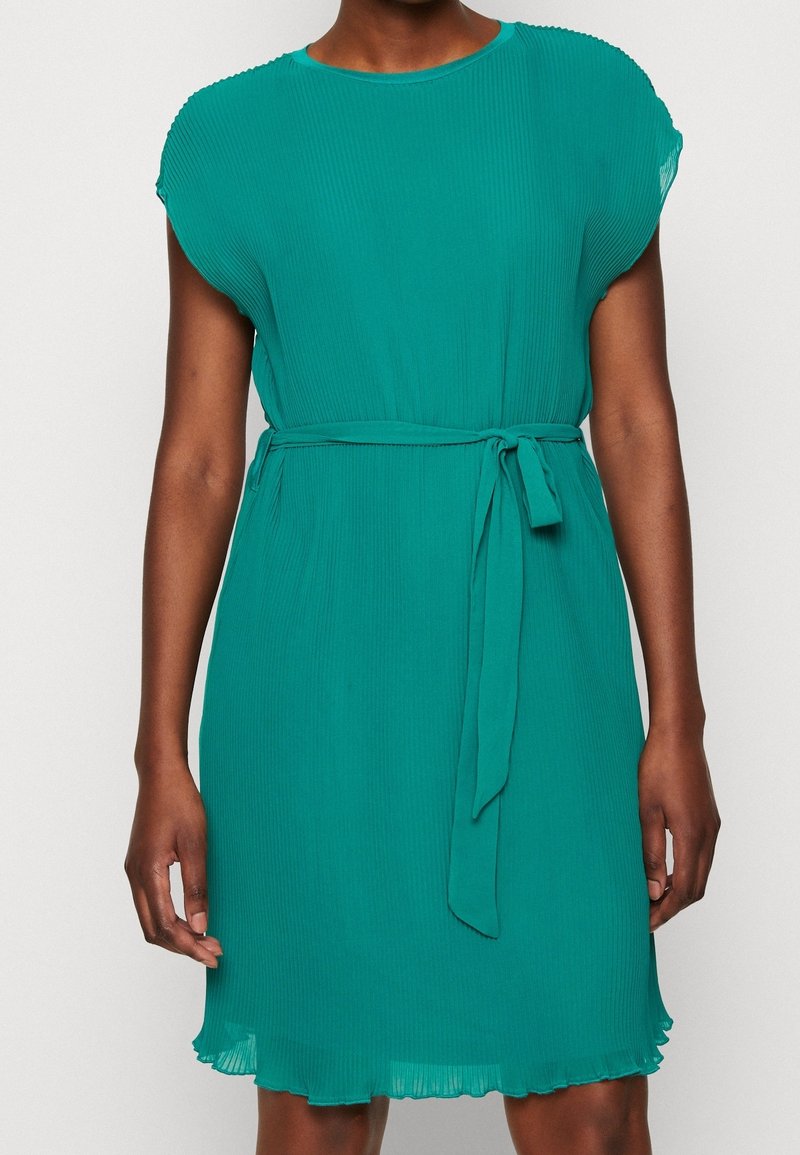 Tealfärgad plisserad klänning med korta ärmar, rund halsringning och ett tygbälte. Har en utsvängd fåll och subtil struktur.