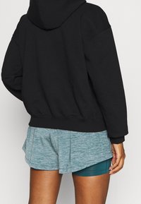Schwarzer Kapuzenpullover kombiniert mit hellen tealfarbenen Sportshorts. Der Pullover hat einen lässigen Schnitt und gerippte Bündchen.