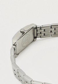 Ezüst rozsdamentes acél karóra téglalap alakú tokban, texturált szíjjal és látható koronaelemmel. Gravírozott márkajelzés a hátoldalán.