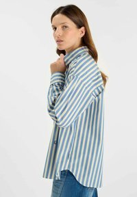 Camicia a righe blu e panna con colletto alto, maniche lunghe e vestibilità ampia. Presenta spacco laterale e orlo curvo per facilitare i movimenti.