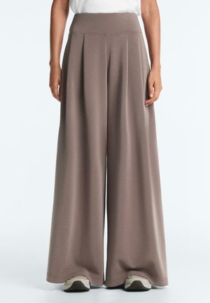 Spodnie o szerokich nogawkach w kolorze taupe z wysokim stanem, noszone z białą bluzką i beżowymi trampkami na jasnym tle.