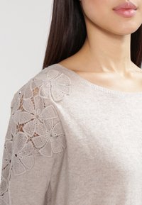 Beige Strickpullover mit transparenter Spitzenblumenverzierung an der Schulter, rundem Ausschnitt und einer weichen Textur.