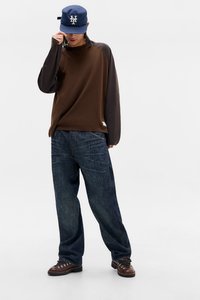 Άτομο που φοράει μπλε καπέλο μπέιζμπολ της ΝΥ, καφέ και σκούρο γκρι μακρυμάνικο πουκάμισο, φαρδιά σκούρα μπλε τζιν και καφέ παπούτσια πεζοπορίας, στέκεται σε λευκό φόντο.