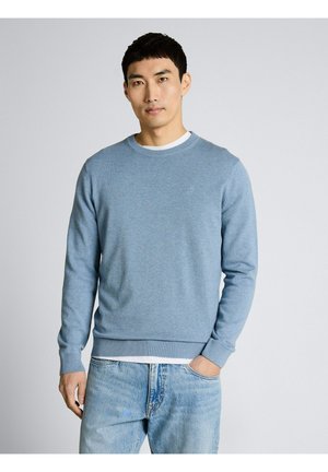 Młody mężczyzna ubrany w jasnoniebieski sweter z okrągłym dekoltem na białą koszulę oraz jasnoniebieskie dżinsy, stojący na tle jednolitego tła, z jedną ręką w kieszeni.