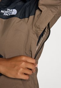 Hnedá a čierna páperová bunda na zips z textúrovanej látky, s kontrastnou panelou a logom, s rukou, ktorá uchopila zips.