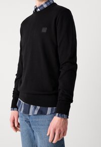Homme portant un pull noir col rond sur une chemise à carreaux bleue et un jean bleu clair, debout devant un fond clair uni.