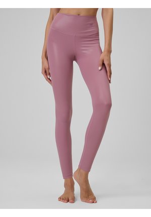Nogi osoby noszącej wysokie, w kolorze mauve legginsy, stojącej boso na jednolitym jasnoszarym tle.