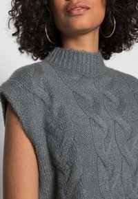 Harmaa korkeakauluksinen neulottu hihaton villapaita, jossa on punoskuvio ja pehmeä koostumus. Yksityiskohdat sisältävät resorikauluksen ja käsiaukot.