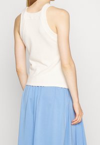 Top in tessuto bianco con scollo rotondo e spalline larghe, abbinato a una gonna plissettata di un azzurro chiaro. La vista posteriore mostra la vestibilità e i sovrapposti.