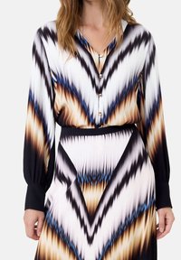Frau trägt ein langärmliges Kleid mit V-Ausschnitt, Knopfleiste und abstraktem Chevron-Muster in Schwarz, Weiß, Braun und blauen Tönen.