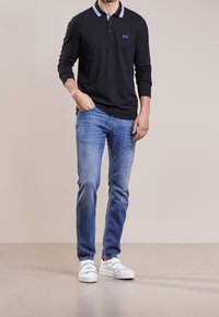 Black long-sleeve polo shirt with striped collar, paired with light blue jeans and white sneakers, standing against a beige background.