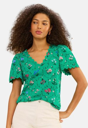 Mujer con cabello rizado, con blusa verde de flores con borde en forma de ondas y pantalones beige, posando con una mano en la cadera, mirando hacia la izquierda.