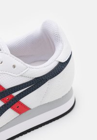 Valkoiset urheilukengät, joissa on verkkokudos ja nahka. Ominaisuuksia punaiset ja laivastonsiniset yksityiskohdat, harmaa välikenkävarsi ja teksturoitu musta ulkopohja.