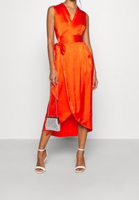 Pomarańczowa satynowa sukienka kopertowa z głębokim dekoltem w serek, wiązana z boku, o długości do kolan. Do tego mała torebka ozdobiona srebrnymi ćwiekami oraz białe szpilki.