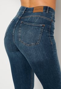 Tmavě modré džíny z denimu s přiléhavým střihem, s hnědou koženou štítkovou páskou a klasickými zadními kapsami, vykazujícími minimální vyblednutí.