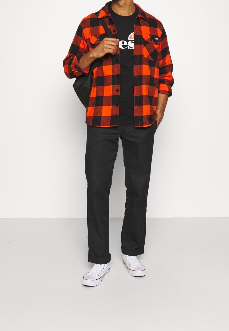 Chemise en flanelle à carreaux rouges et noirs portée sur un t-shirt noir, associée à un pantalon foncé et des baskets blanches, avec un sac à bandoulière.