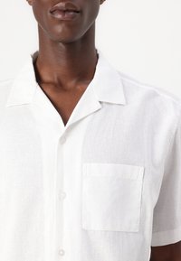 Short-sleeved white linen shirt with a notch collar, single chest pocket, and button closure. Soft texture and lightweight design.