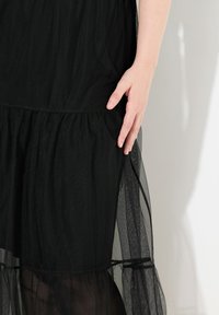 Schwarzes Kleid aus strukturiertem Stoff, mit einem geschichteten Tüllrock und einem elastischen Taillenbund. Eine Hand ruht auf dem Stoff in der Nähe der Taille.