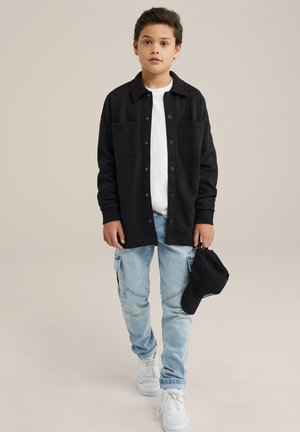 Camicia nera a maniche lunghe con bottoni sopra una t-shirt bianca, jeans cargo azzurri chiari e sneakers bianche. Cappellino nero tenuto nella mano destra.