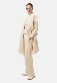 Beżowy oversize'owy szal z frędzlami, zestawiony z dopasowanym kremowym golfem i szerokimi kremowymi spodniami. Całość dopełniają sportowe białe buty.