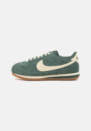 Baskets basses en daim vert avec un logo Nike swoosh crème, des lacets crème et une semelle en gomme à motif strié.