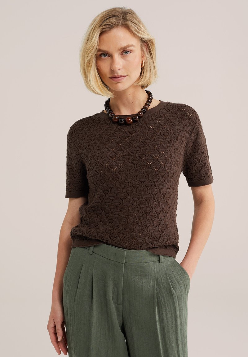 Femme blonde portant un haut à manches courtes marron foncé texturé, un collier épais de perles foncées, et un pantalon taille haute plissé vert olive.