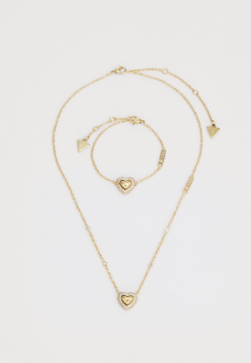 L'ensemble de bijoux en or comprend un collier et un bracelet, ornés de pendentifs en forme de cœur avec des accents en pierre claire et de charms en forme de triangle avec le logo GUESS.