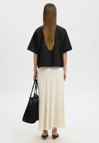 Haut court noir avec finition en dentelle, associé à une longue jupe crème. Le mannequin tient un sac fourre-tout noir texturé. Cheveux bruns lisses et raides visibles.