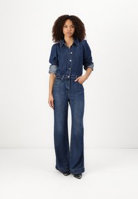Denim jumpsuit med lange ermer, knappestolpe foran og belted midje. Mørkeblå farge, vid ben-design og kontrasterende lyseblå mansjetter.