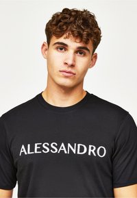 Musta puuvillapaita, jossa on valkoisella langalla brodeerattu "ALLESSANDRO" teksti rintakehässä. Klassinen pyöreä kaula-aukko ja lyhyet hihat.