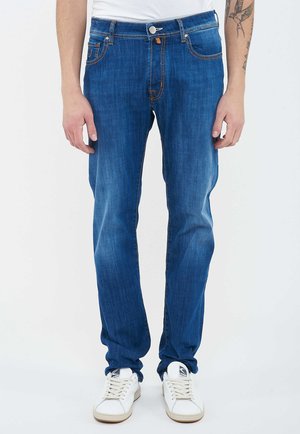 Man wearing medium blue slim-fit jeans with faded details and white sneakers, standing against a white background.