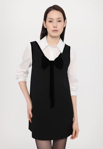 Zwarte mouwloze jurk met een fluwelen strik bij de halslijn, gelaagd over een witte blouse, gecombineerd met zwarte panty's. Gladde stof textuur.