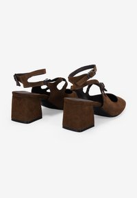 Zapatos de tacón cuadrado de ante marrón con diseño recortado, correas ajustables en el tobillo y punta redonda. El tacón grueso proporciona estabilidad.