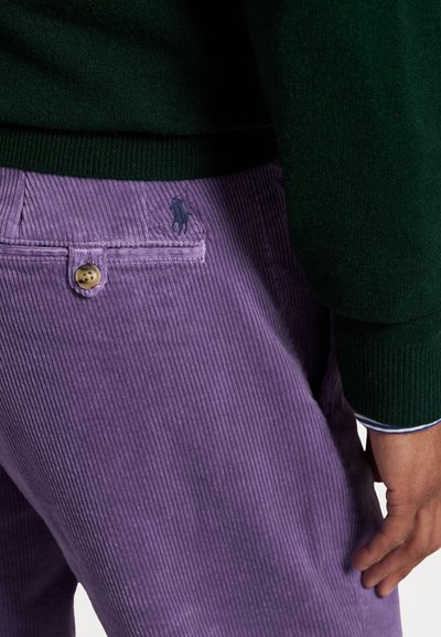 Paarse corduroy broek heeft verticale ribbels, een achterzak met een knoop en een klein geborduurd logo. Gecombineerd met een donkergroene trui.