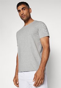 Grå kortærmet t-shirt i et blødt, bomuldsagtigt materiale med tre hvide chevron-detaljer på venstre skulder. Rund halsudskæring, afslappet pasform.