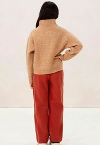 Beiger gerippter Pullover mit hohem Kragen, kombiniert mit roten Jogginghosen mit weißen Seitenstreifen, von hinten betrachtet.