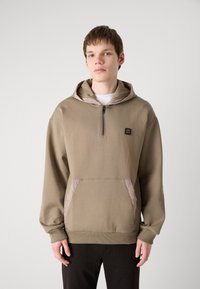 Sudadera con cremallera color caqui y capucha acolchada, que cuenta con un bolsillo frontal y un parche con logo. Textura suave, corte relajado y mangas largas.