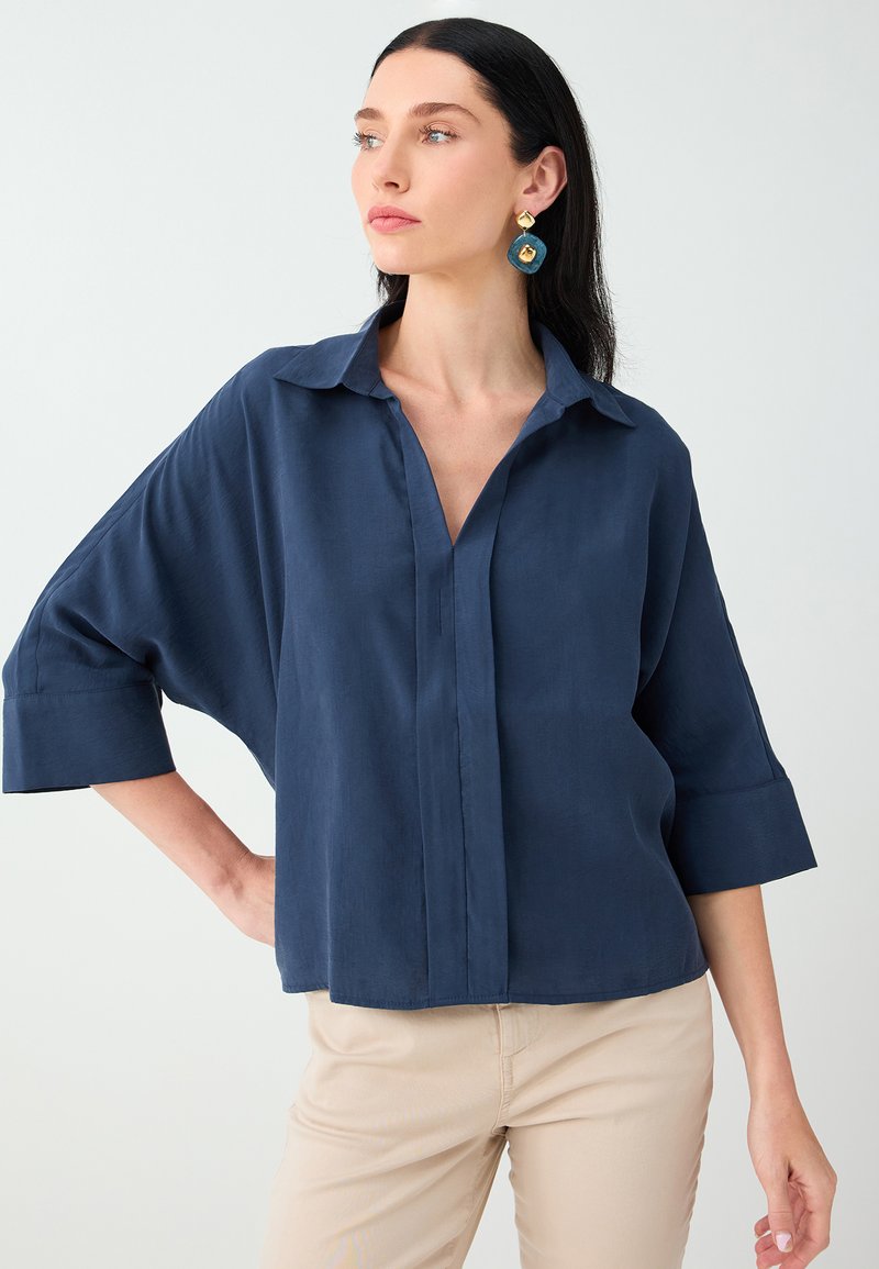Femme aux cheveux foncés portant une chemise bleu marine ample à manches trois-quarts et un pantalon beige, regardant sur le côté devant un fond uni.