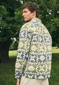 Junger Mann, der im Freien unter einem Baum steht und eine gesteppte Jacke mit grünen, blauen und weißen geometrischen Sternmustern trägt.