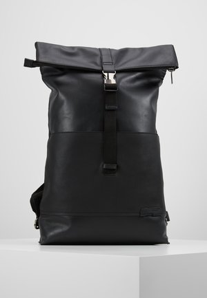 Mochila negra con cierre enrollable, correa con hebilla frontal y cremallera, textura de cuero suave, sobre una superficie blanca frente a un fondo gris claro.