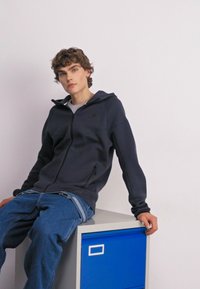 Schwarze Zip-Up-Hoodie mit strukturierter Oberfläche, ausgestattet mit einer Kapuze und seitlichen Taschen. Kombiniert mit blauen Jeans. Auf einem grauen Aufbewahrungselement sitzend.